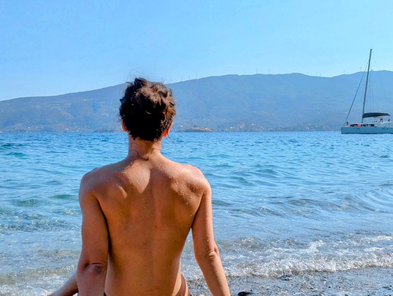 Person seen from behind, sitting at the shoreline and visible from the waist up, with a clearly built upper body, broad shoulders and a strong back, facing the sea. Calm blue water, distant mountains, and a sailboat to the right under a clear sky.