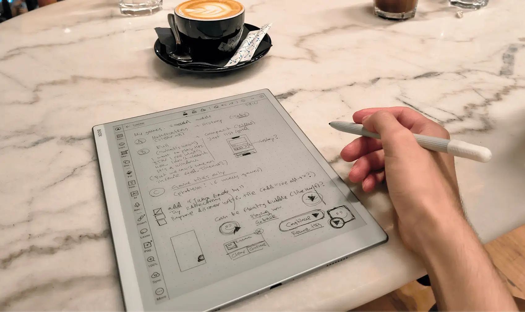 A tablet with an E-Paper screen on a table in a cafe. Latte visible in the background. Hand visible holding a Wacom pen.